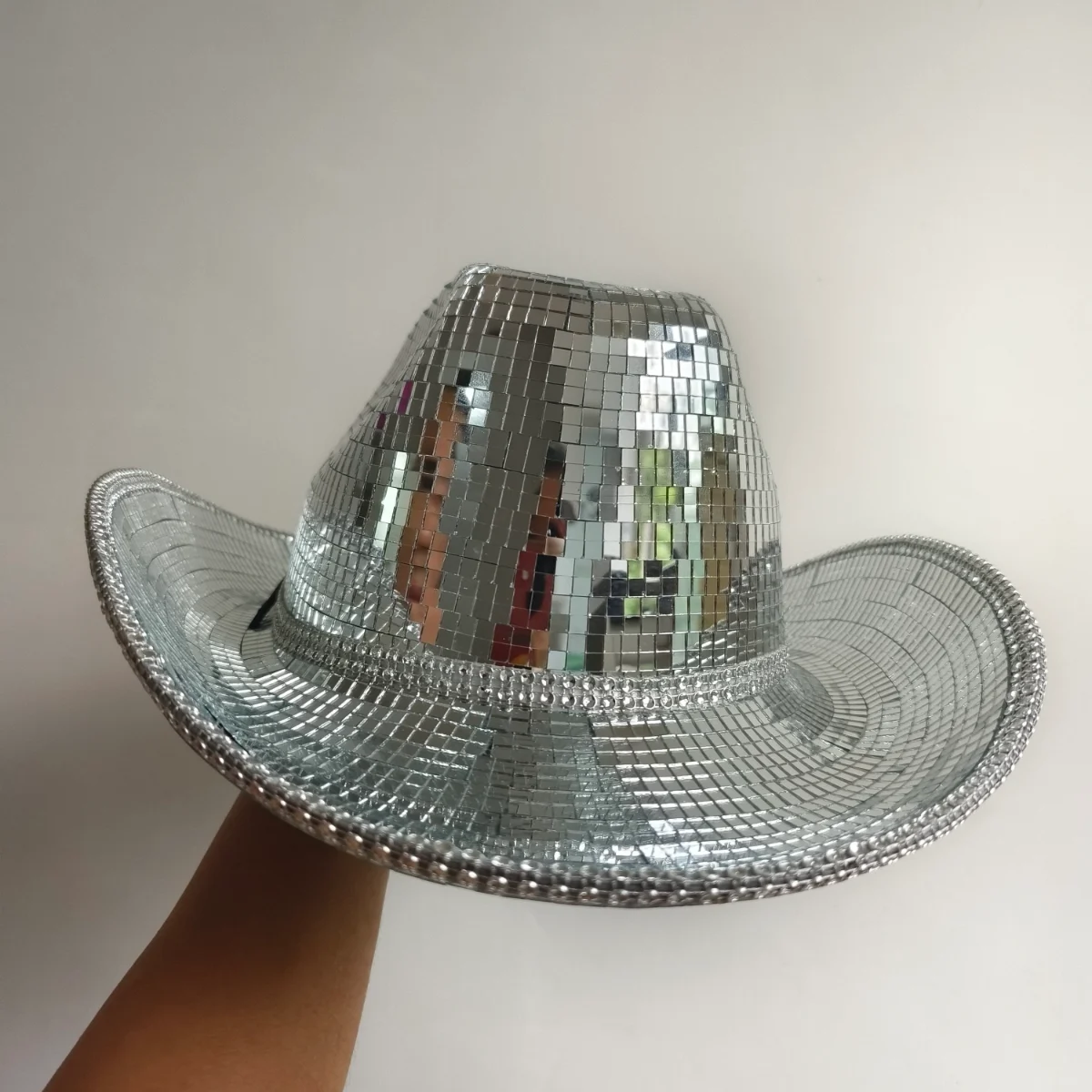 Hand holding a silver disco ball mirror cowboy hat against a plain wall